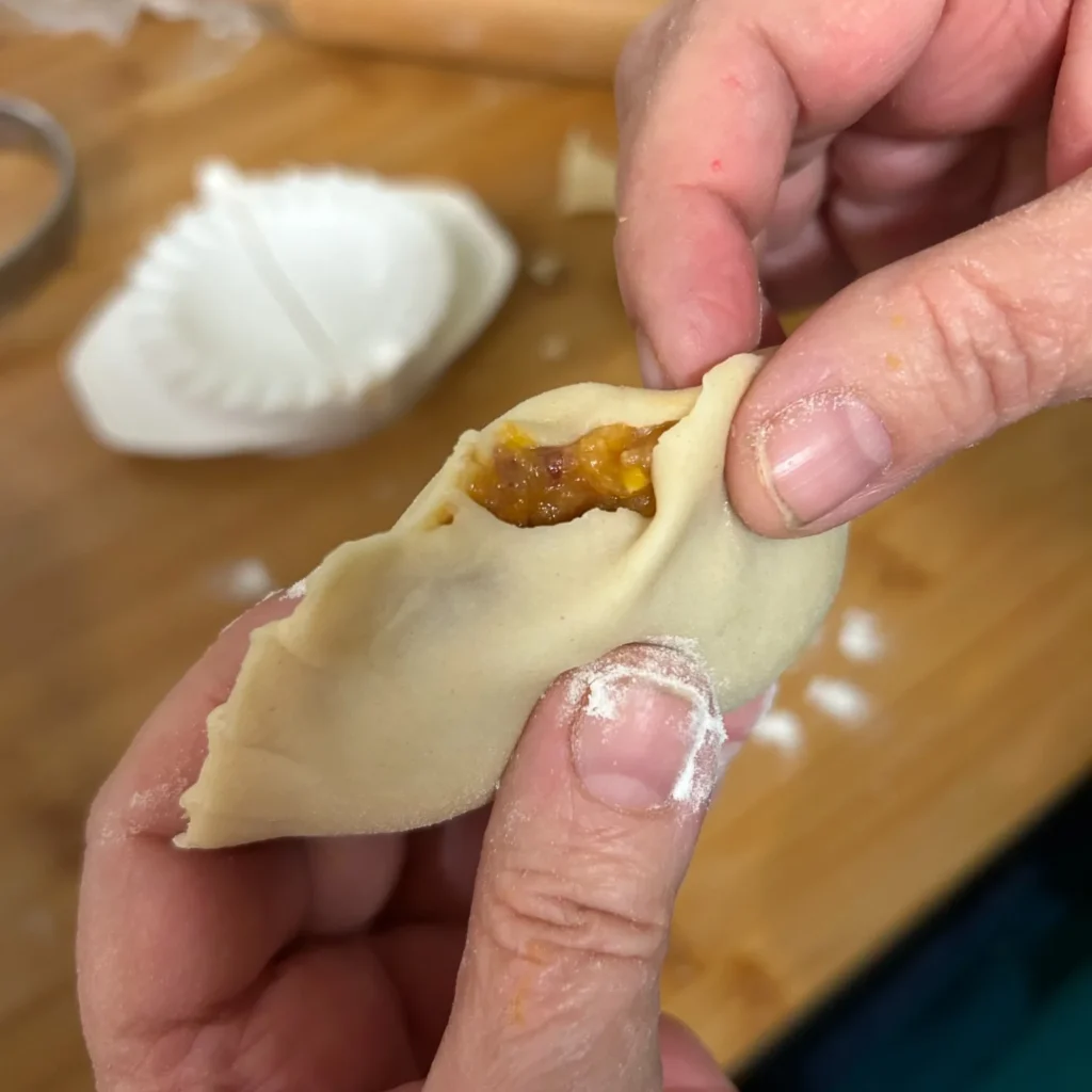 Hands pinching a pierogi closed with a glimpse of orange Pilgrim's Harvest natural filling on a floured work surface.