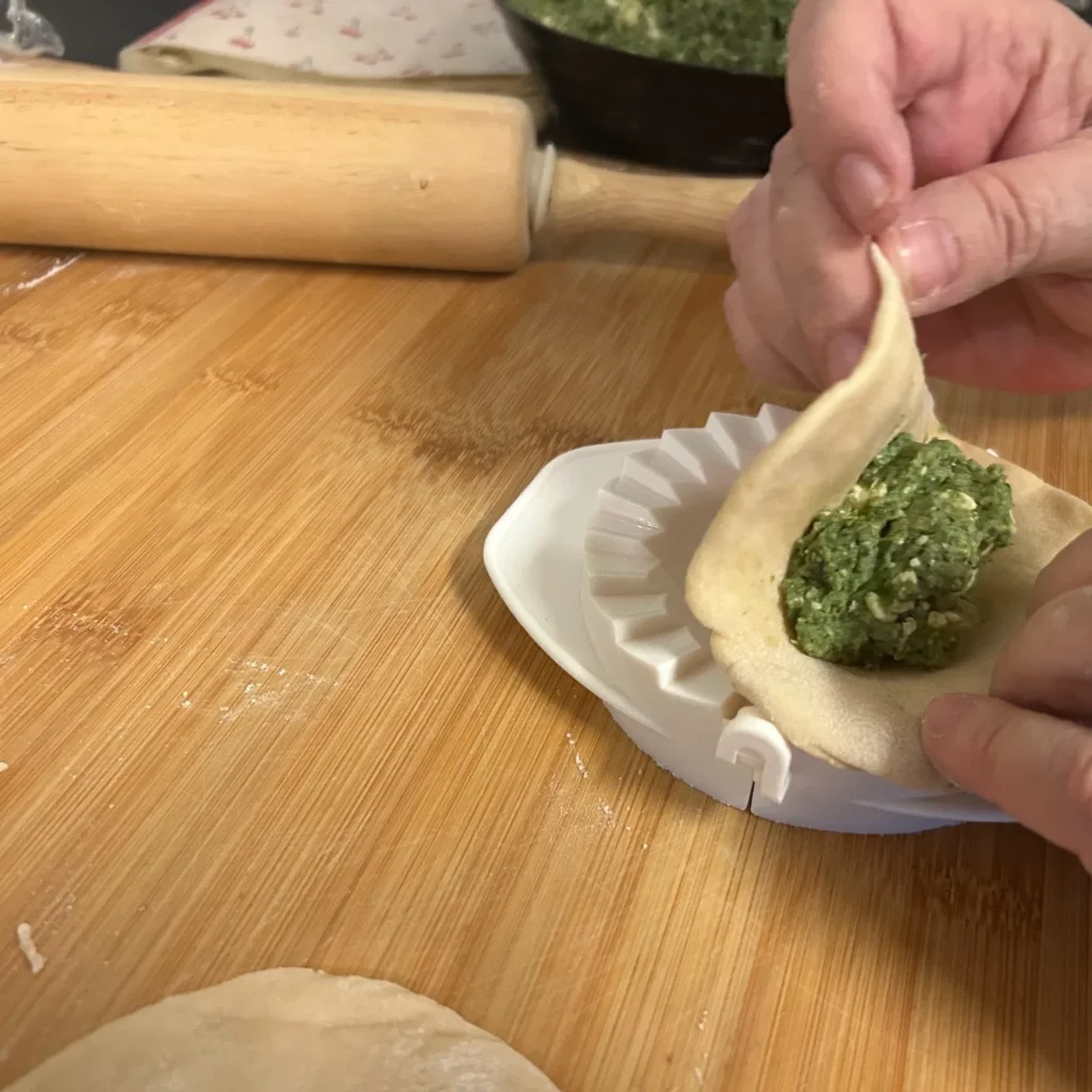 Hands folding dough around spinach and feta filling on a single pierogi press with a rolling pin nearby on top of a wood grain prep board.