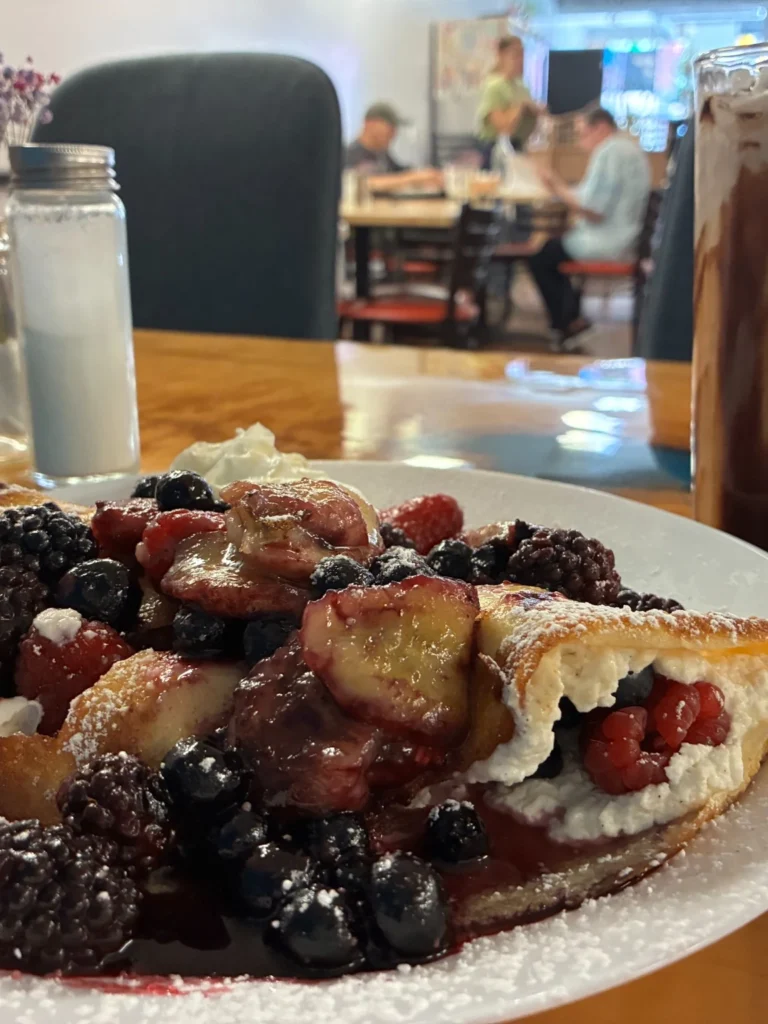 Loaded fruit blintz-style sweet crepe piled with mixed berries, whipped cream, powdered sugar, and berry sauce beside an iced coffee drink.