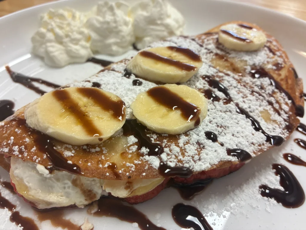 Vanilla sweet cheese banana crepe blintz dusted with powdered sugar, topped with banana slices, chocolate drizzle, and whipped cream.