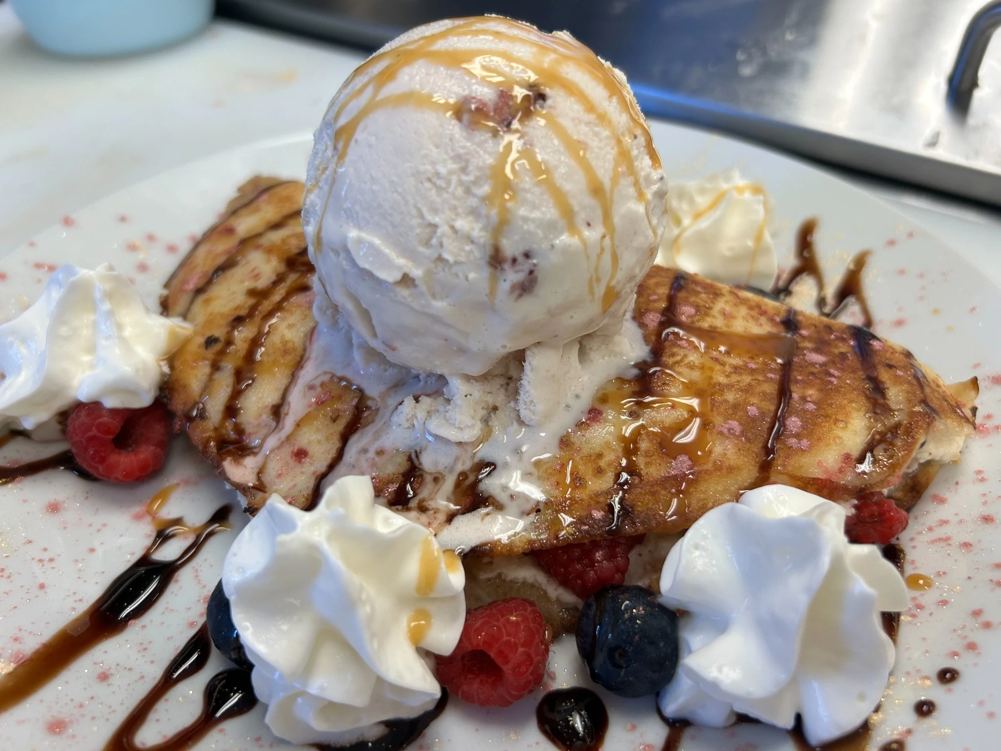 Dessert crepe with vanilla sweet cheese, berries, whipped cream, and a large scoop of vanilla ice cream melting over caramel drizzle.