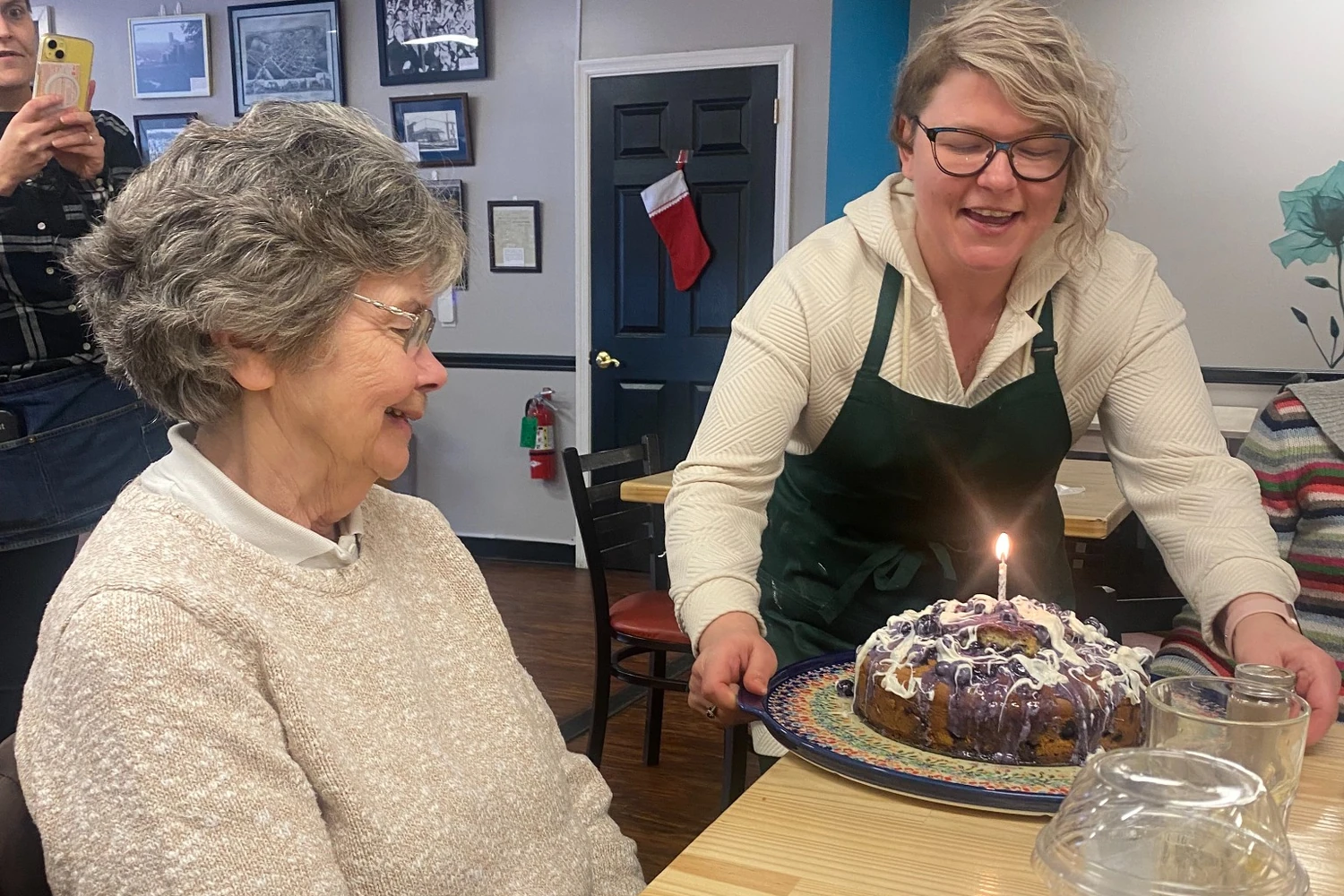 Chef Kat carrying a lit birthday dessert cake to a smiling guest during a celebration inside the café-restaurant.