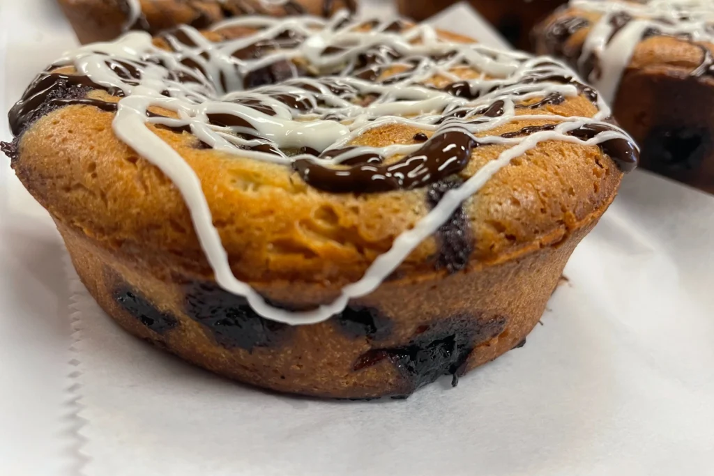 Blueberry chocolate muffin mini cakes topped with white icing and chocolate drizzle in the bakery case.