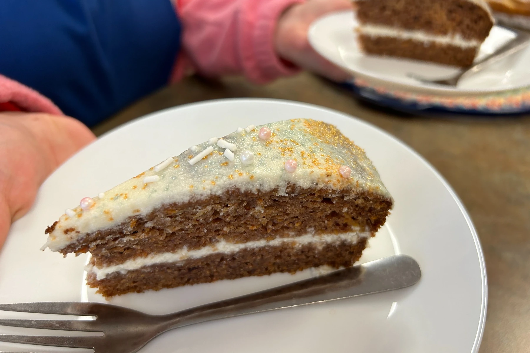 Slice of carrot cake with cream cheese frosting and festive sugar pearls on a white plate with a fork.