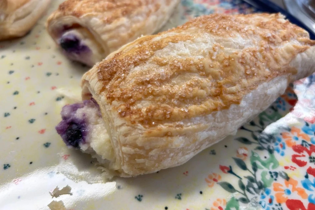 Flaky cheese and berry croissant pastries with creamy filling and blueberries peeking from the ends on a floral plate.