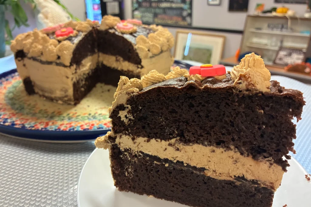 Slice of dark chocolate cake layered with coffee cream frosting, with the full decorated cake in the background.