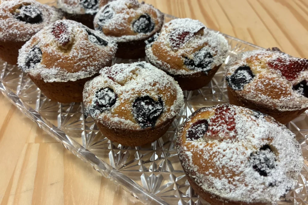 Mini berry muffins studded with blueberries and strawberries, dusted with powdered sugar and arranged on a crystal tray.