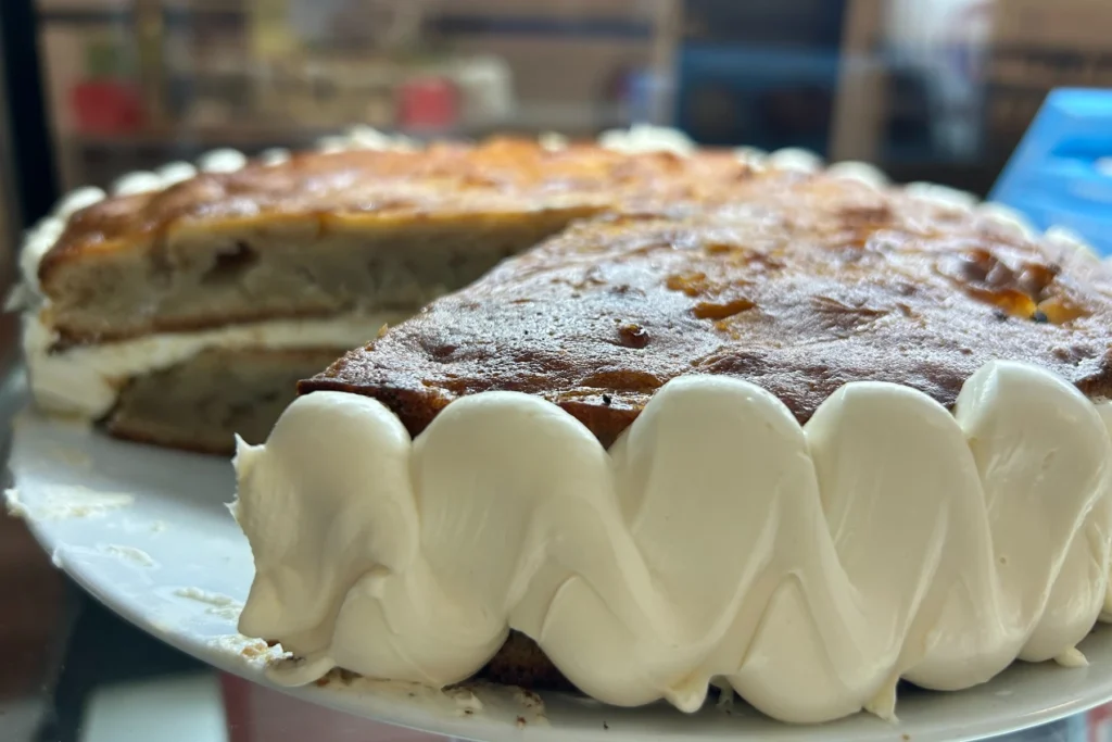 Fresh baked layered sponge cream cake with a slice removed, showing cream filling and thick piped frosting around the sides.