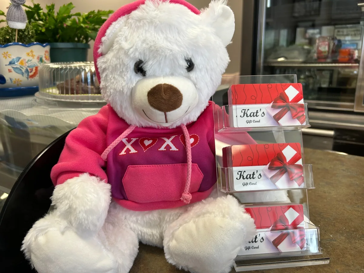 White teddy bear in a pink hoodie next to a display of Kat’s Café & Restaurant gift cards.