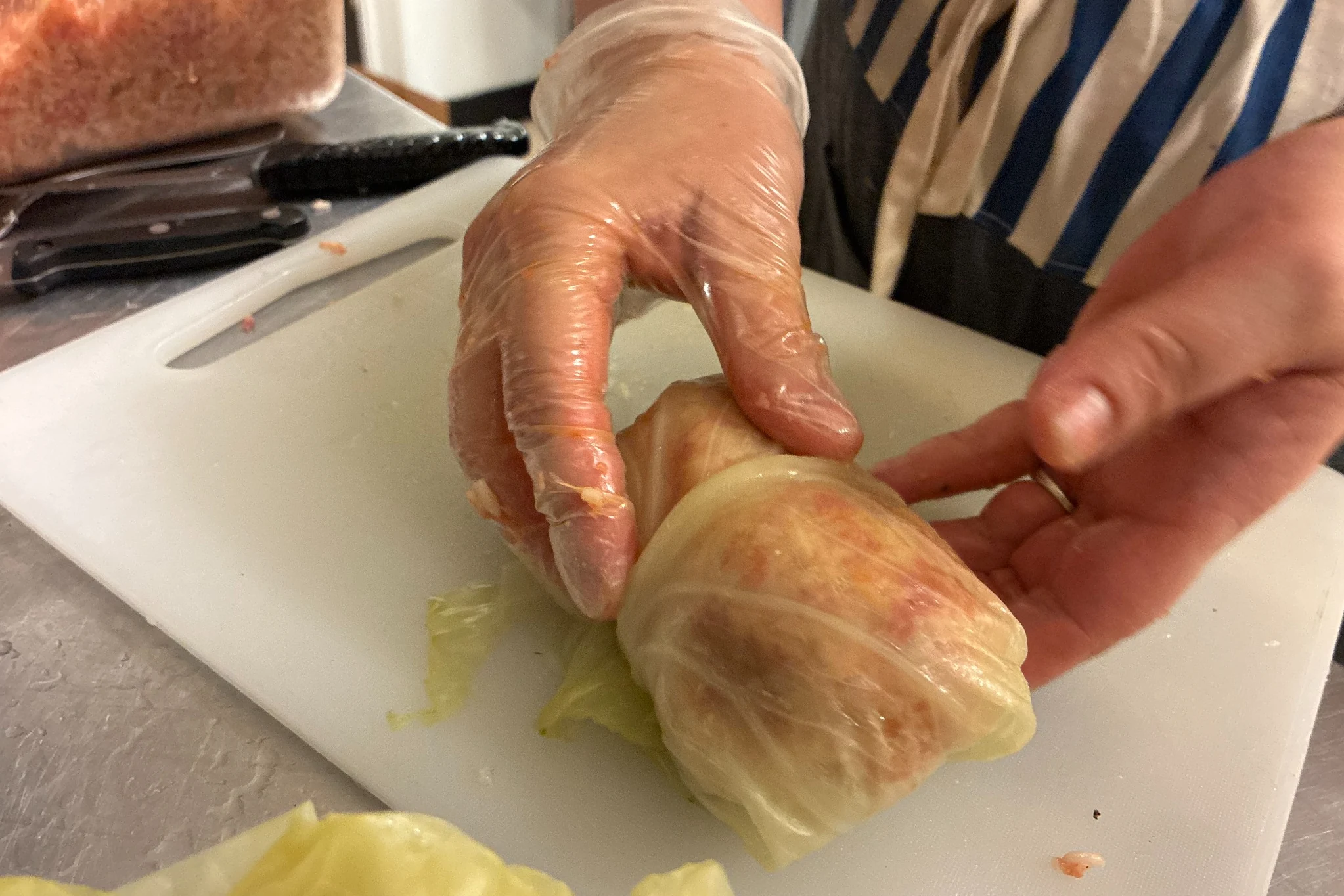 Chef Kat's gloved hands rolling a fresh halupki in a cabbage leaf on a prep board.