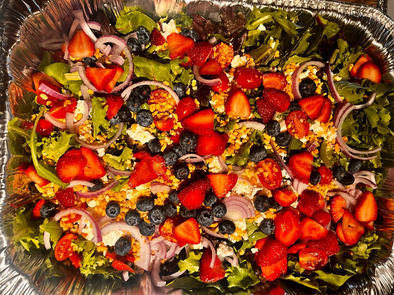 Catering salad tray with mixed greens, strawberries, blueberries, tomatoes, red onion, cheese, and chopped nuts.