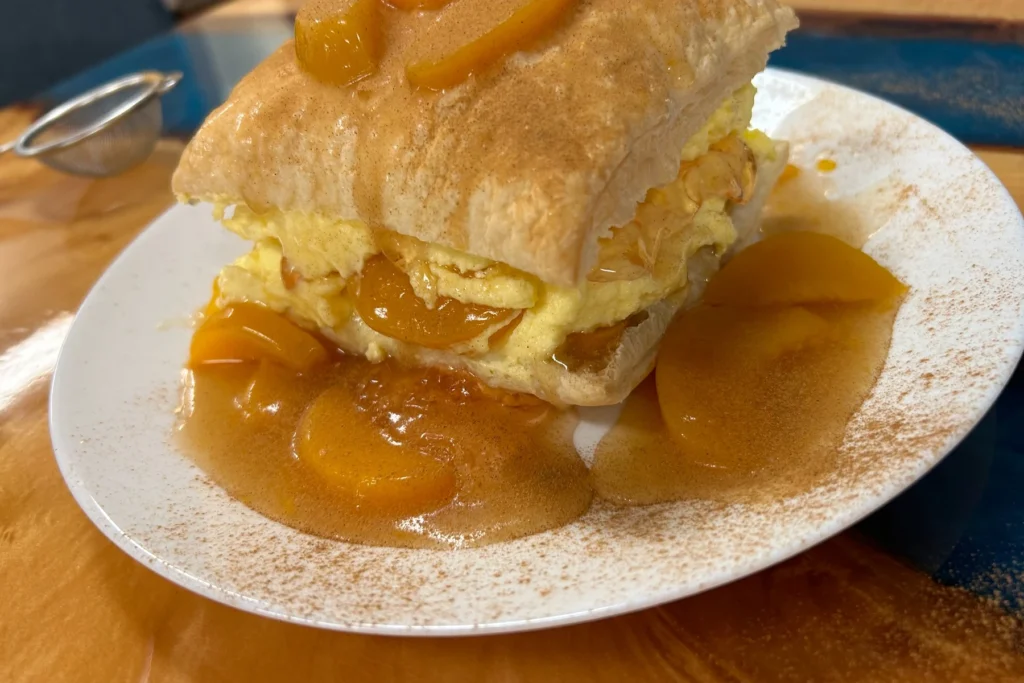 Peach Slavic Napoleon pastry layered with real cream and peach filling, plated with warm fruit sauce and a dusting of cinnamon.