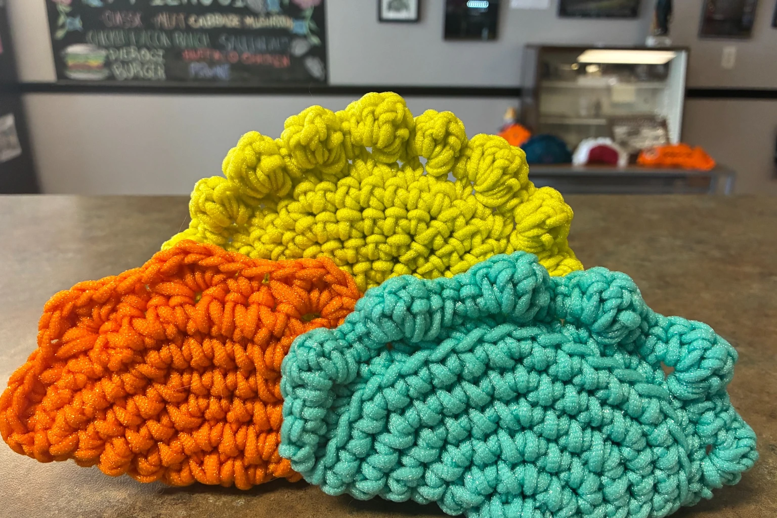 Colorful crochet scrubbies at Kat’s Three colorful handmade crochet scrubbies displayed on the counter at Kat’s Café & Restaurant with the pierogi chalkboard softly blurred in the background.