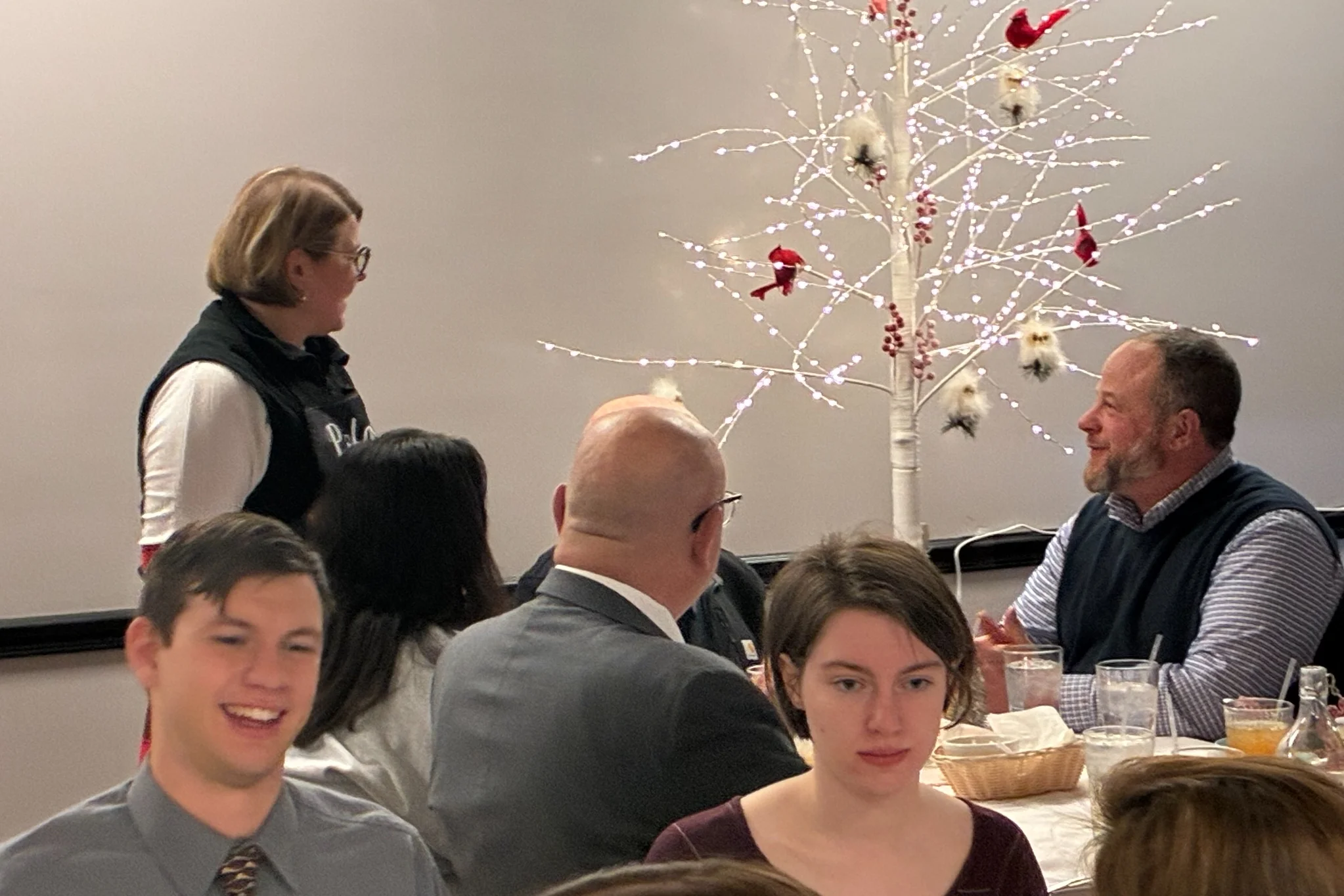 Guests listening Chef Kat speaks beside a lit white holiday tree during a Christmas Eve Holy Supper at Kat’s Café & Restaurant.