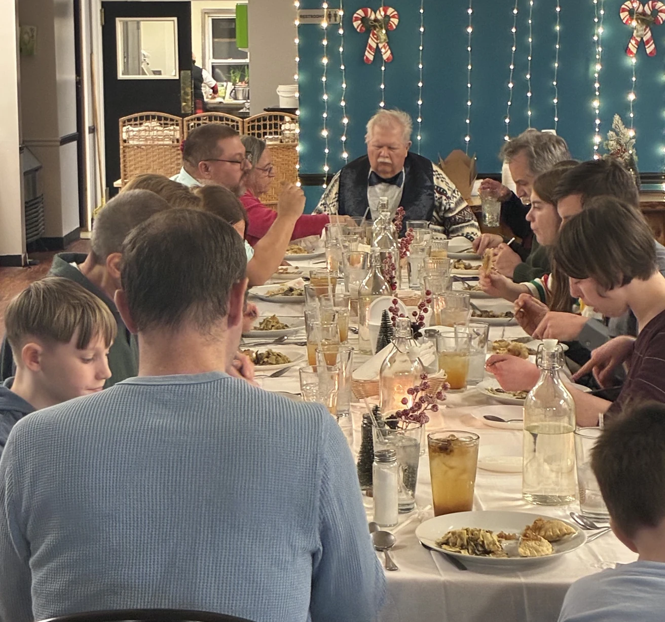 Guests seated around a long table during Kat’s Café & Restaurant Christmas Eve Holy Supper, with holiday lights in the background.