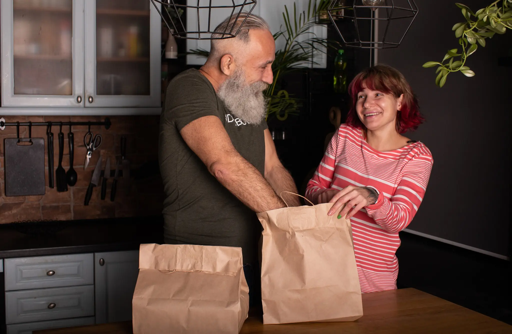 Two smiling customers unpacking brown paper takeout bags in a home kitchen.