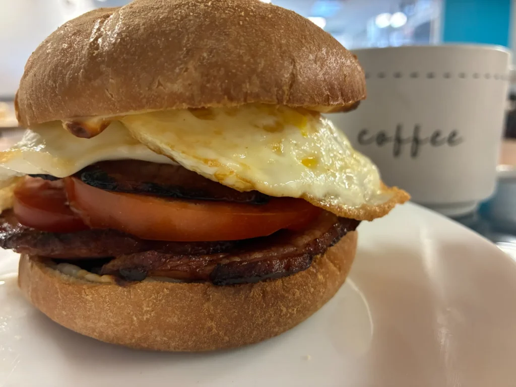 Breakfast sandwich with grilled ham, tomato, melted cheese, and fried eggs beside a coffee mug at Kat’s Café & Restaurant.