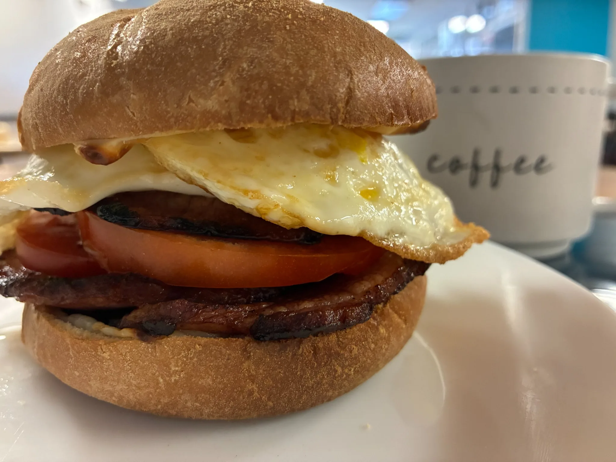 Breakfast sandwich with grilled ham, tomato, melted cheese, and fried eggs beside a coffee mug at Kat’s Café & Restaurant.