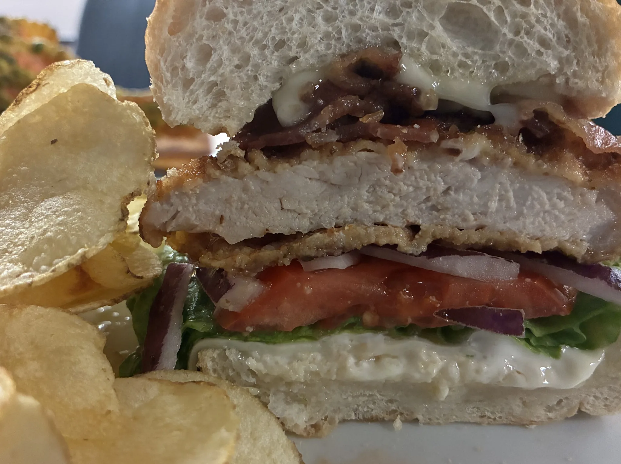 Close-up of a pork schnitzel sub with bacon, melted cheese, lettuce, tomato, red onion, and creamy mayo, served with potato chips at Kat’s Café & Restaurant.