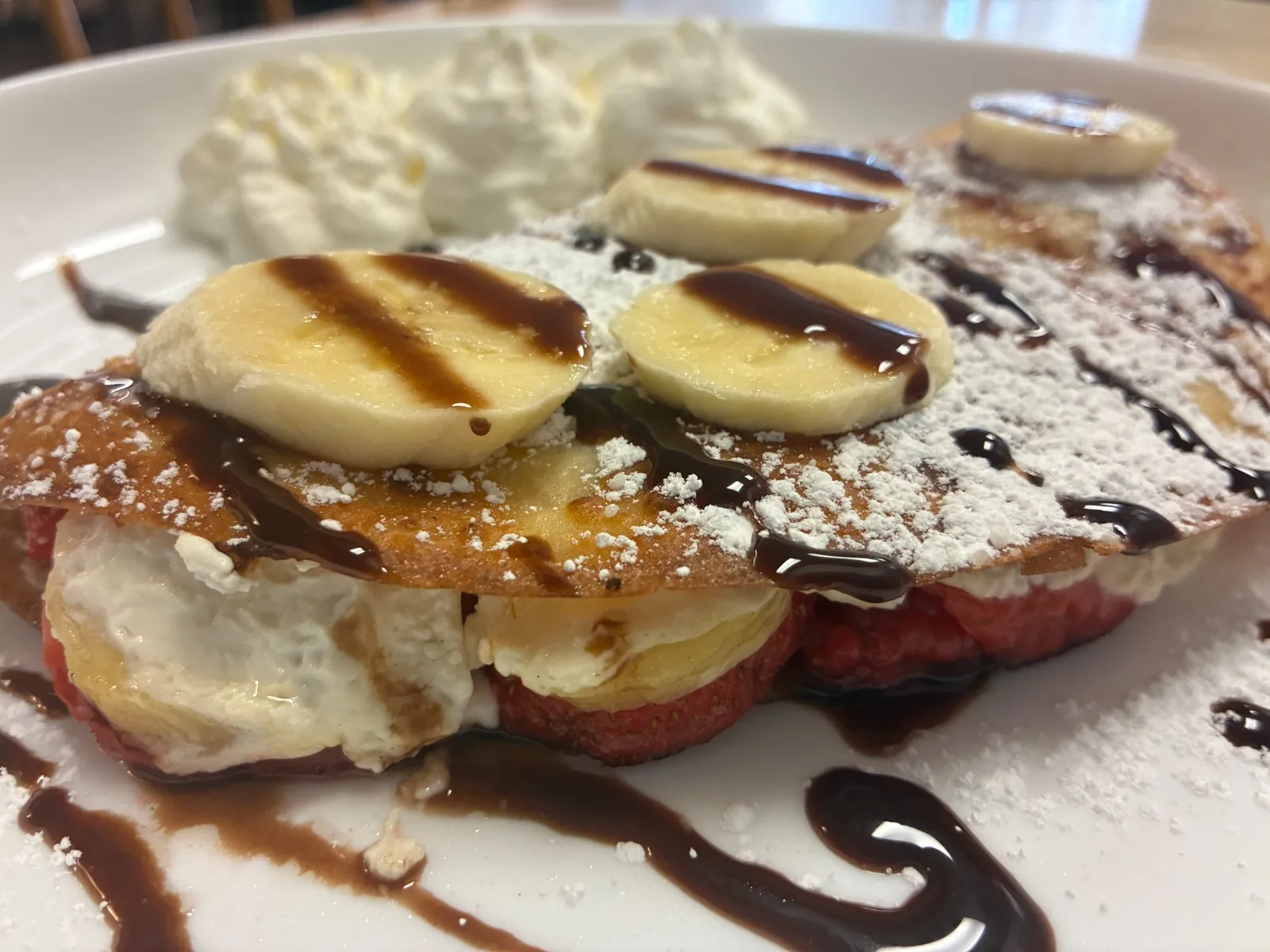 Folded sweet cheese crepe with strawberries and banana, topped with powdered sugar and chocolate drizzle and served with whipped cream served at Kat’s Café & Restaurant.