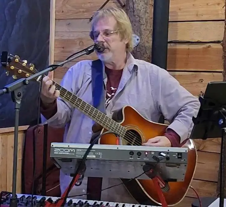 Local musician John Derek singing and playing acoustic guitar with a keyboard in front of him during a live performance.