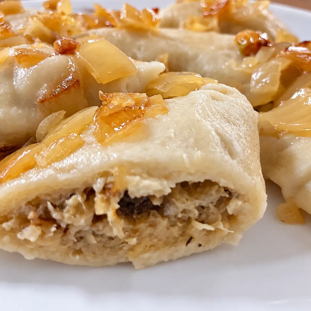 Handmade cabbage and mushroom pierogies topped with buttery sautéed onions, with one cut open to show the house-made filling.