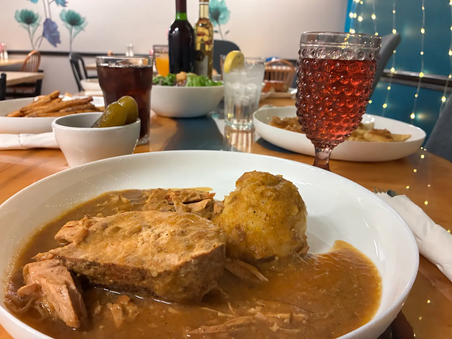 A hearty roast dinner covered in savory brown gravy fills the foreground of a table set with red and white wine bottles, cold drinks, additional plates, and a decorative ruby-colored goblet at Kat’s Café & Restaurant.