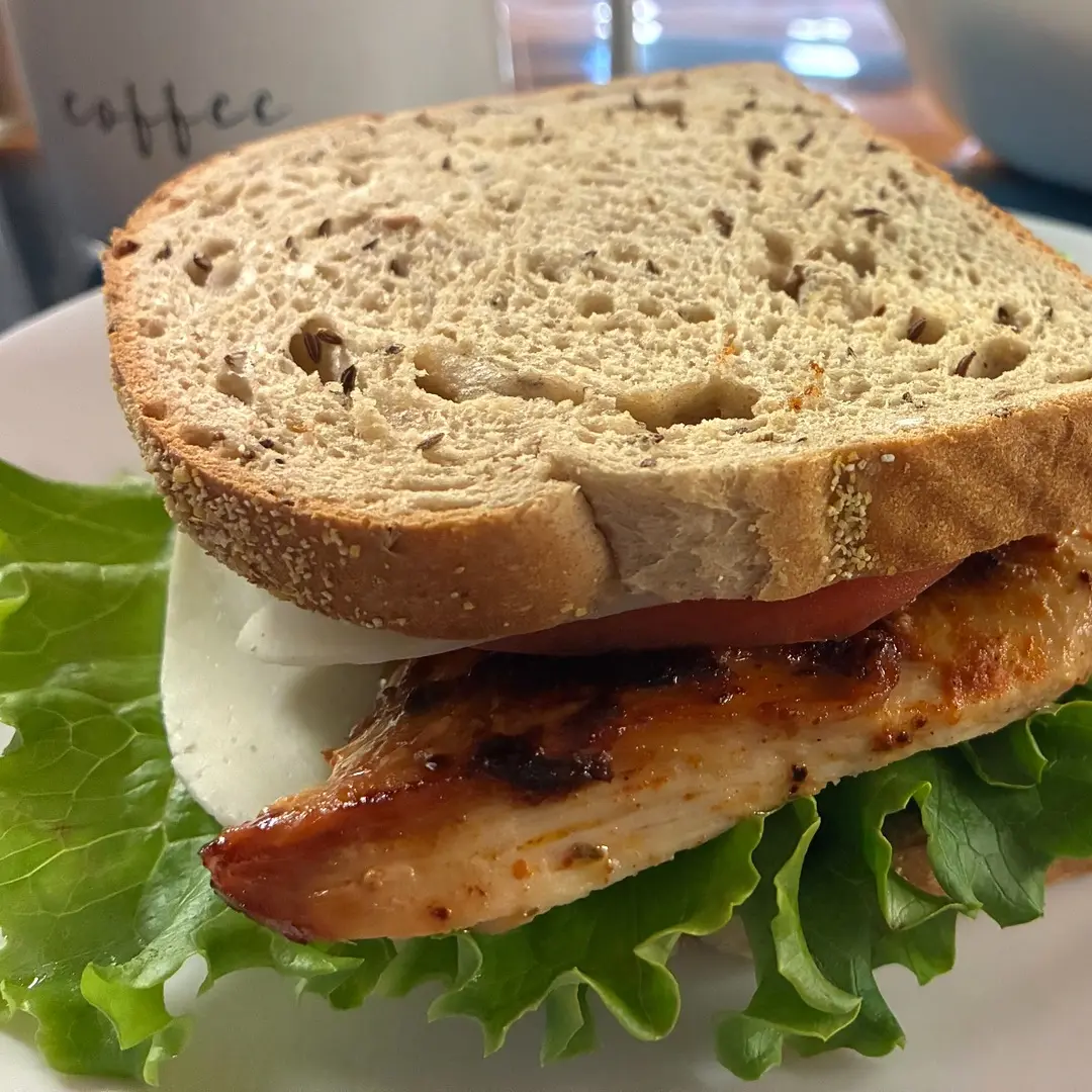 Grilled chicken breast sandwich at Kat’s Café & Restaurant with lettuce, tomato, and white cheese on seeded bread, served on a white plate.