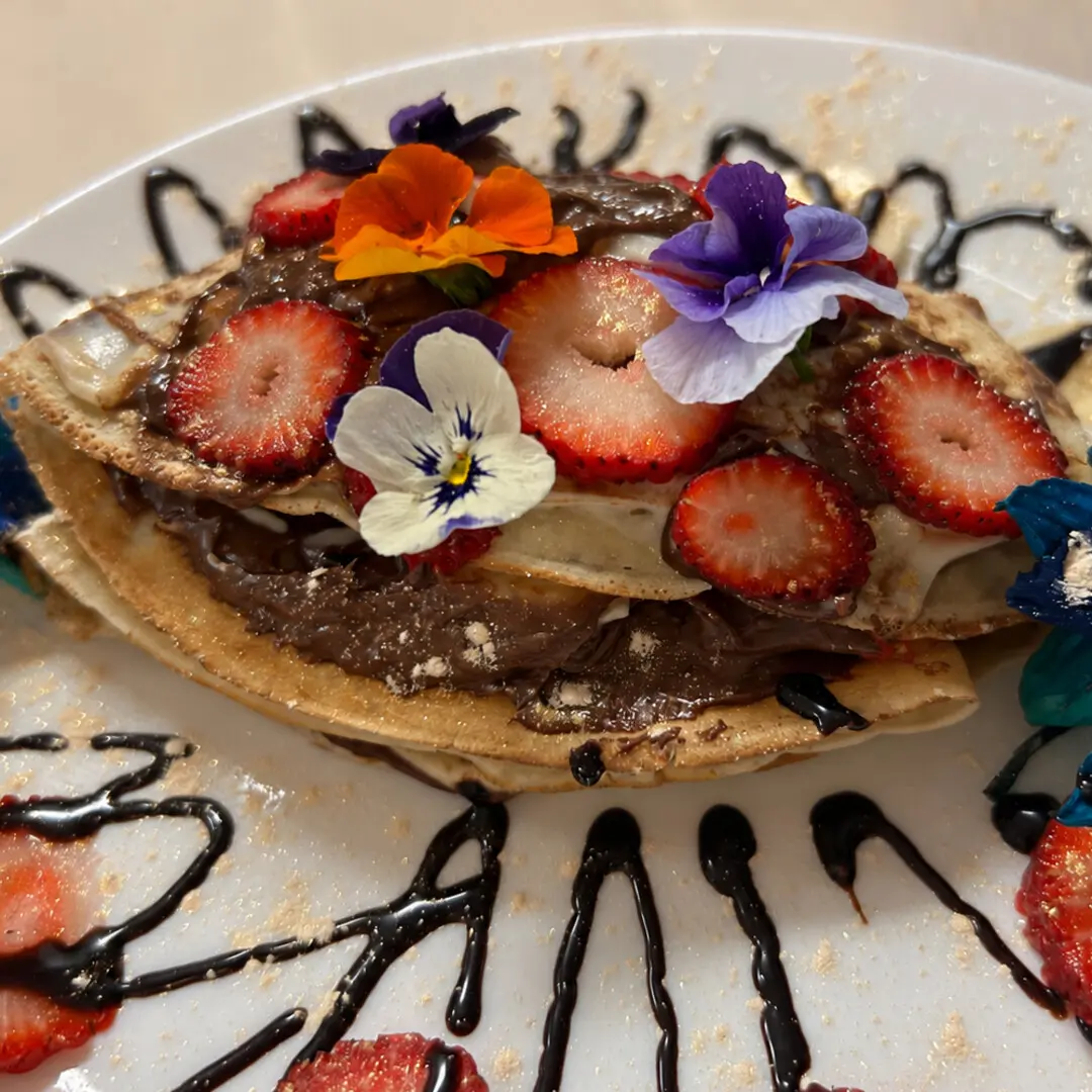 Freshly made crepe layered with Nutella and fresh strawberries, topped with more strawberries, powdered sugar, edible flowers, and chocolate drizzle.