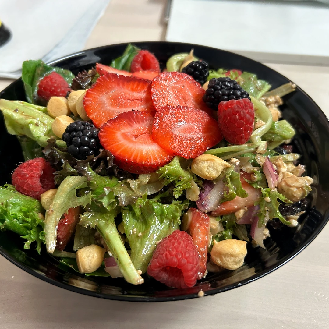 Kat’s Nuts n Berries House Salad with mixed greens, strawberries, raspberries, blackberries, nuts, tomatoes, and red onion in a black bowl.
