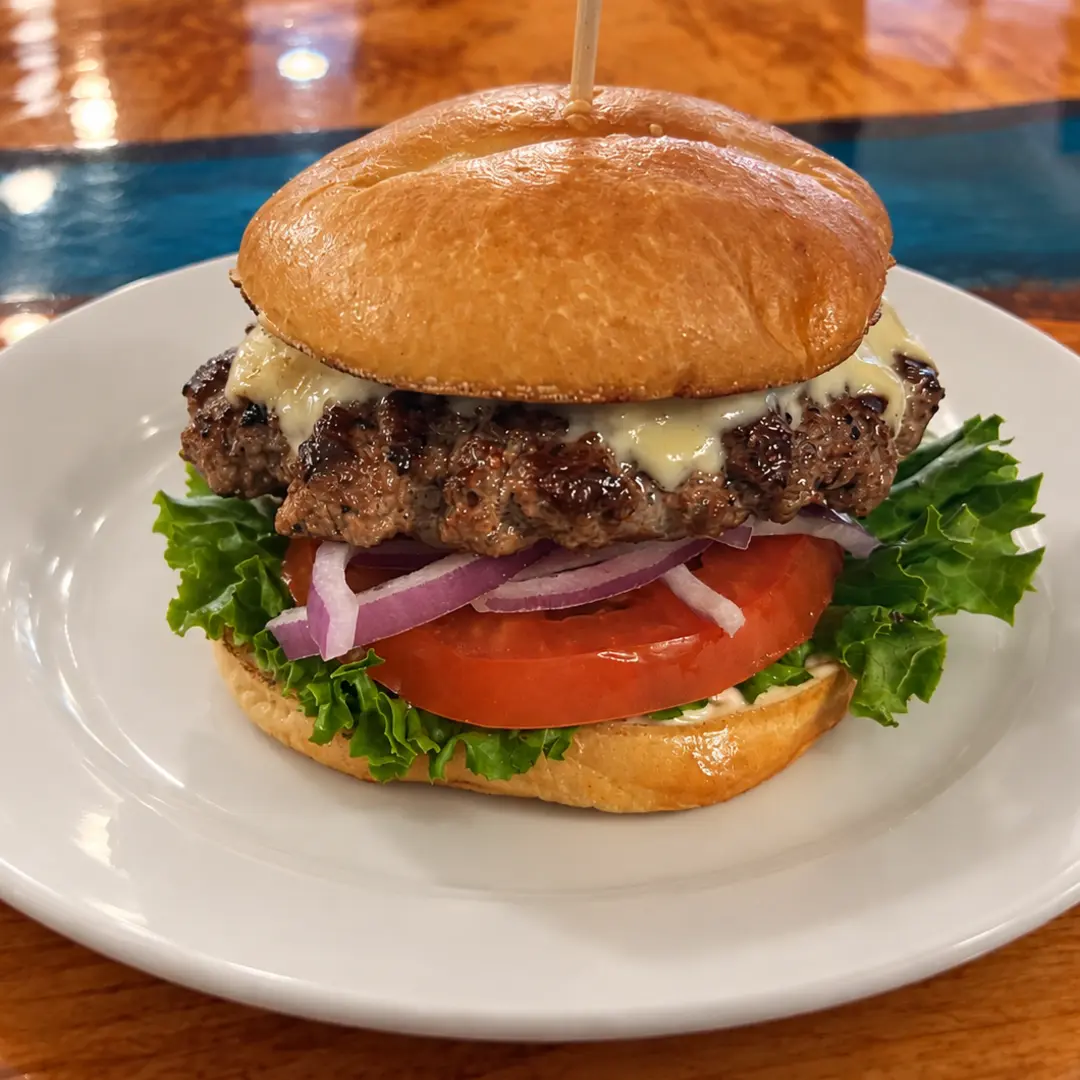 Smash burger at Kat’s Café & Restaurant with melted cheese, lettuce, tomato, and red onion, served on a white plate atop the custom blue-resin river dining room table.