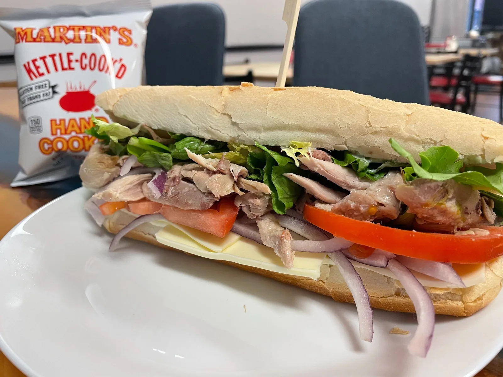 Grilled chicken sub at Kat’s Café & Restaurant, with lettuce, tomato, red onion, and sliced cheese on a hoagie roll, served on a white plate with a bag of kettle-cooked chips behind it.