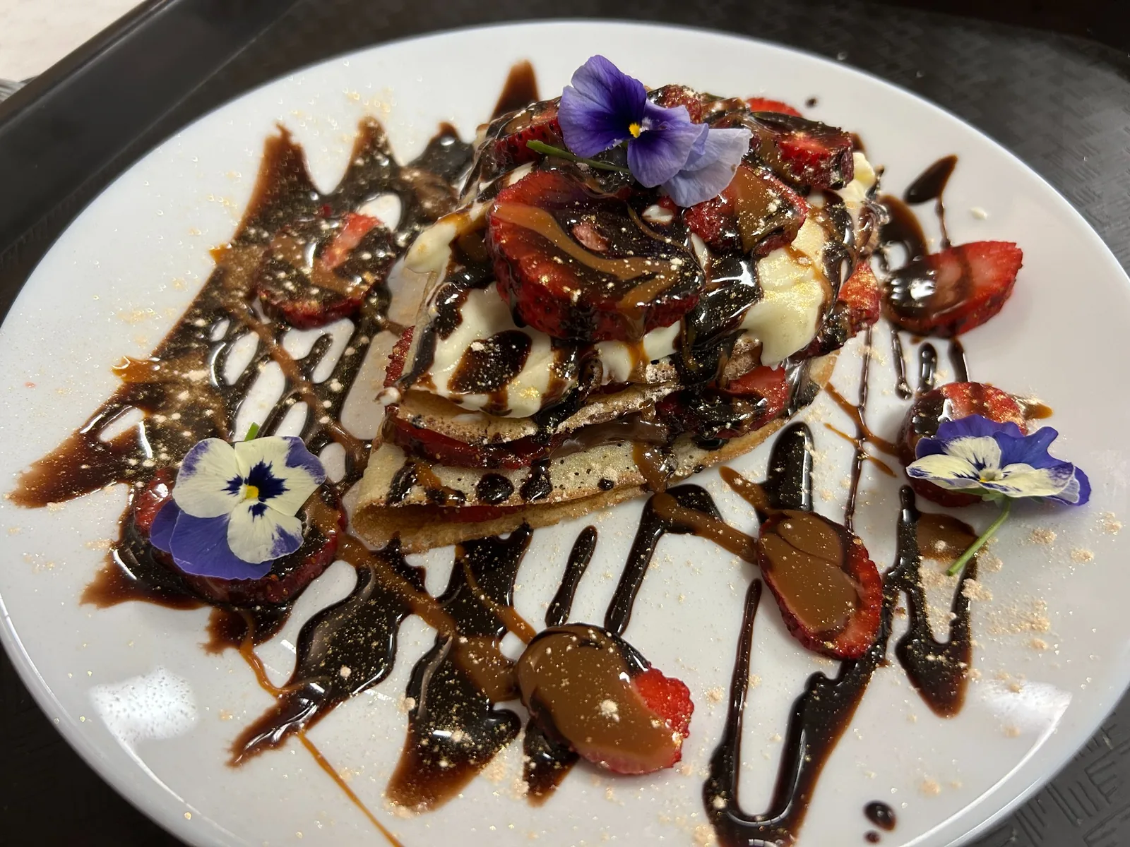 Folded Nutella crepe at Kat’s Café & Restaurant, layered with sliced strawberries and topped with cream, sauce drizzles, and purple flowers on a white plate.